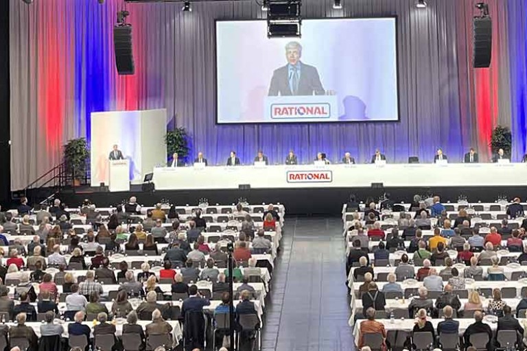 Eine große Menschenmenge bei einer Hauptversammlung in einem Konferenzsaal, mit einem Redner auf der Bühne.