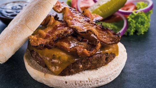 A close-up of a bacon cheeseburger with melted cheese, served with vegetables and dipping sauce.
