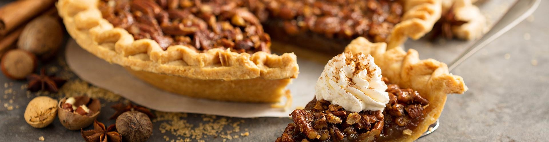 Una torta di noci pecan su un tavolo con una fetta sormontata da panna montata, circondata da zucche e spezie.