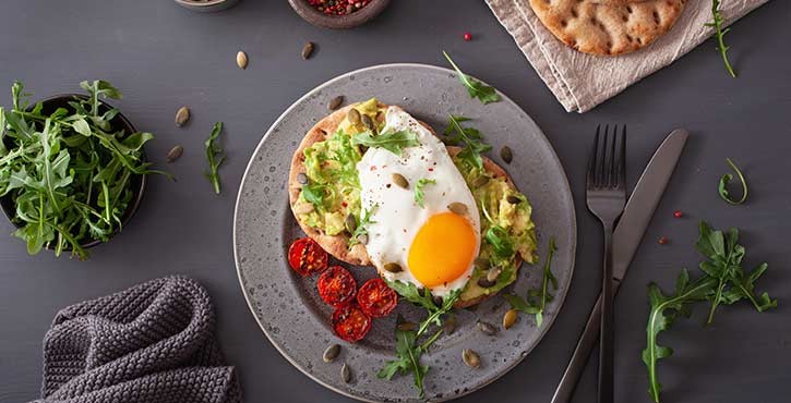 Une assiette de petit-déjeuner gastronomique comprenant un sandwich à l'avocat garni d'un œuf au plat, entouré de roquette et de tomates rôties.
