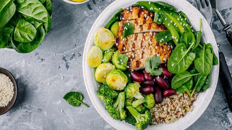 A colorful Buddha bowl featuring grilled chicken, quinoa, broccoli, Brussels sprouts, and fresh greens, garnished with sesame seeds.