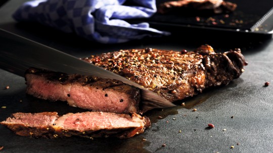 Primo piano di una bistecca di ribeye alla griglia che viene affettata, mettendo in evidenza il succoso interno rosa e il condimento in cima.