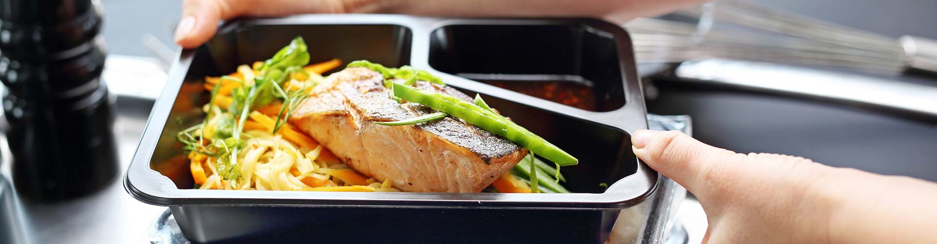 A person holding a stack of meal prep containers with salmon, noodles, and vegetables, ready for delivery or serving.
