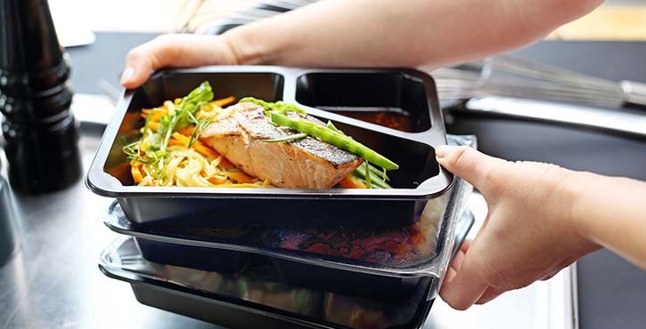 Una persona sosteniendo un montón de recipientes de comida con salmón, fideos y verduras, listos para la entrega o el servicio.