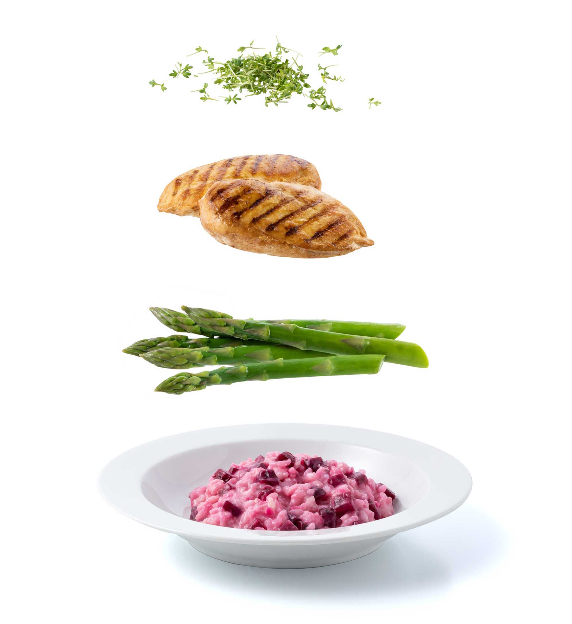 Arrangement artistique de filets de poulet grillés, d'asperges, de thym et d'un bol de risotto à la betterave sur fond blanc.