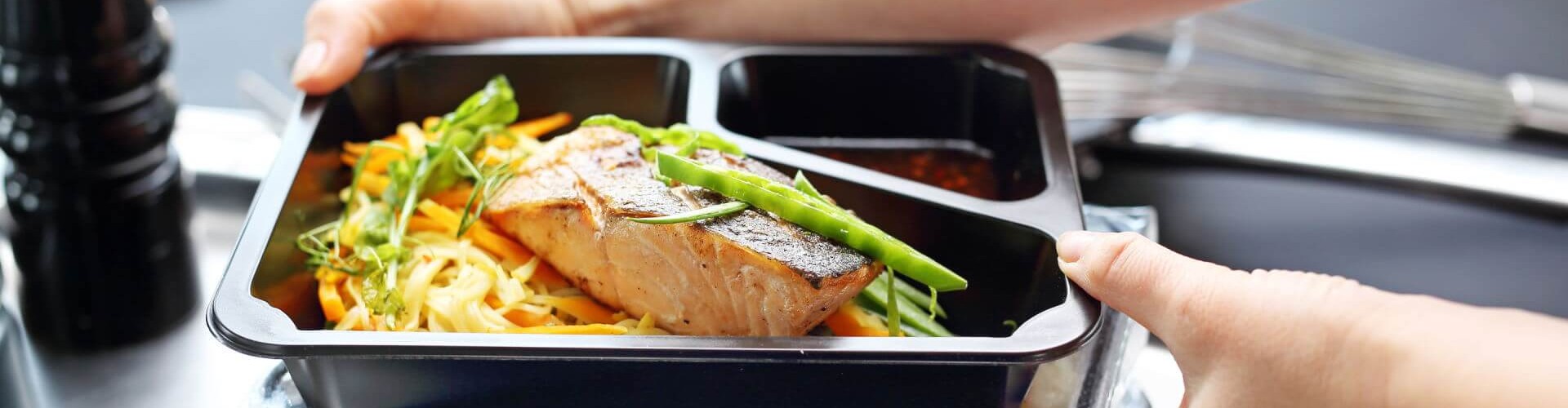 A person holding stacked lunchboxes filled with salmon and vegetables, ready for service.