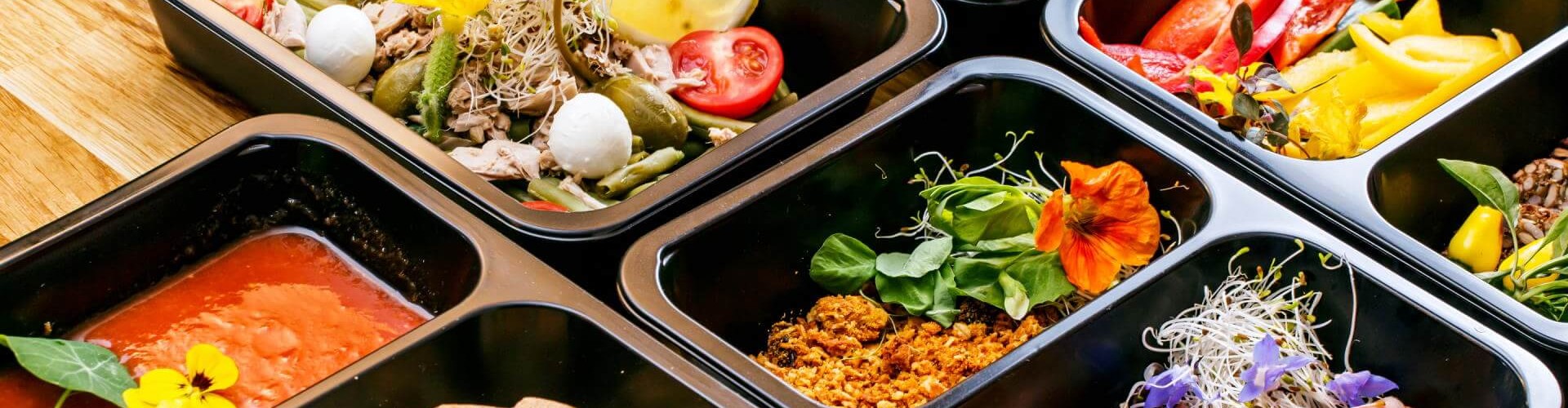A variety of convenience food boxes containing fresh salads, sauces, and sliced meats, beautifully garnished with edible flowers.