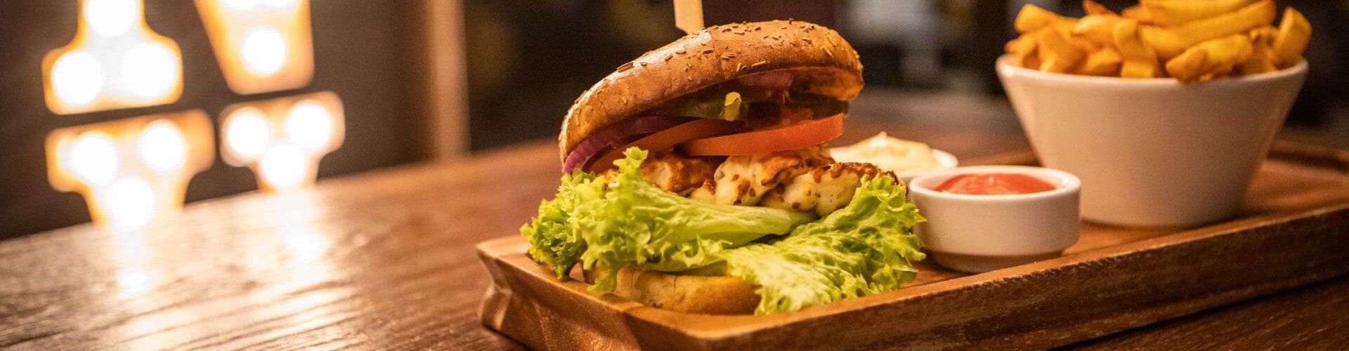 Un hamburger gourmet con lattuga, pomodoro, cipolla e sottaceti servito su un piatto di legno, accompagnato da un contorno di patatine e salse da intingere.