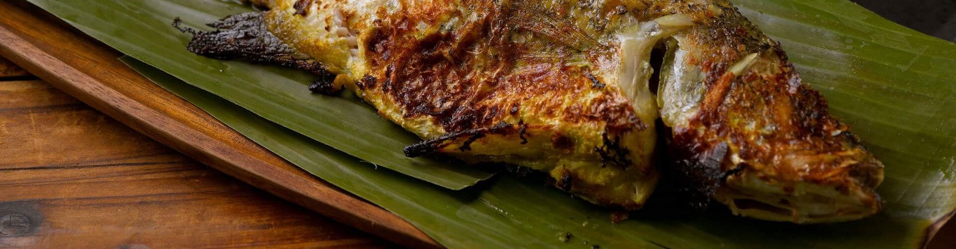 Grilled fish served on banana leaves with three lime halves beside it, showcasing a crispy texture and rich colors.