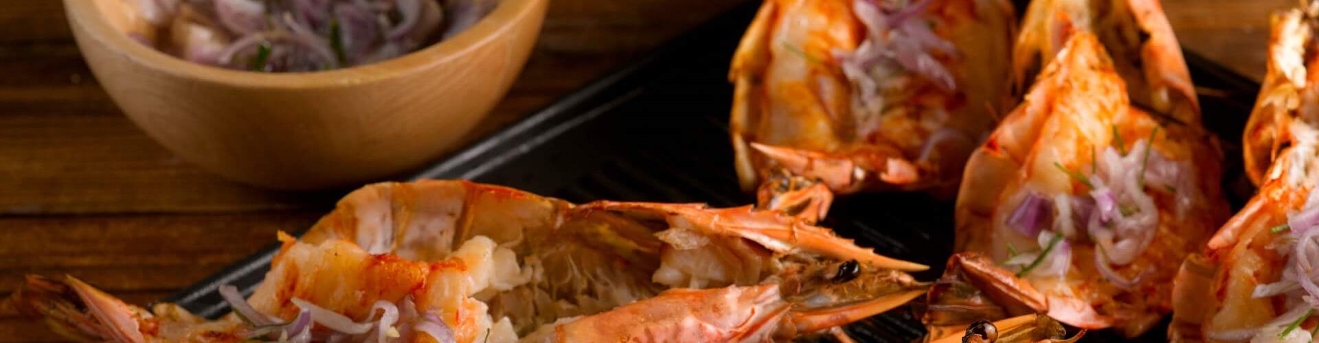 Grilled prawns served on a grill plate with sliced shallots in a wooden bowl, showcasing a rich orange color and a savory presentation.