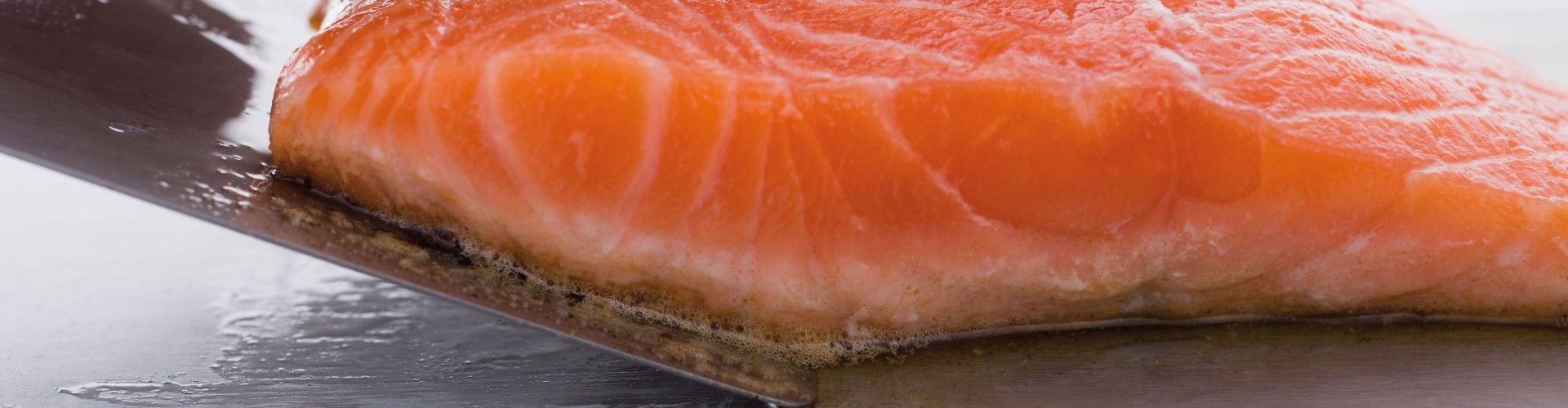 Eine Nahaufnahme einer Hand, die vorsichtig auf ein Stück rohen Lachs auf einer Edelstahloberfläche drückt und dessen Textur und Frische zeigt.
