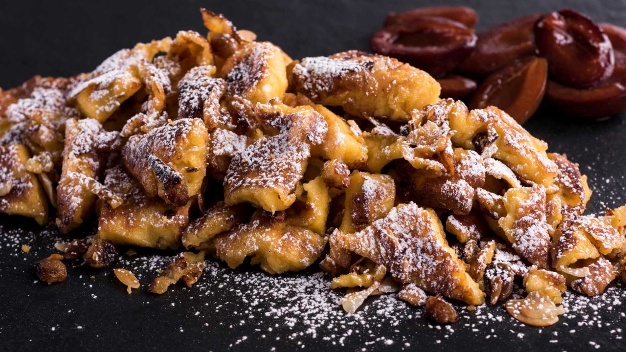 Plate of kaiserschmarrn, a shredded pancake dessert dusted with powdered sugar, served with halved plums on a dark surface.