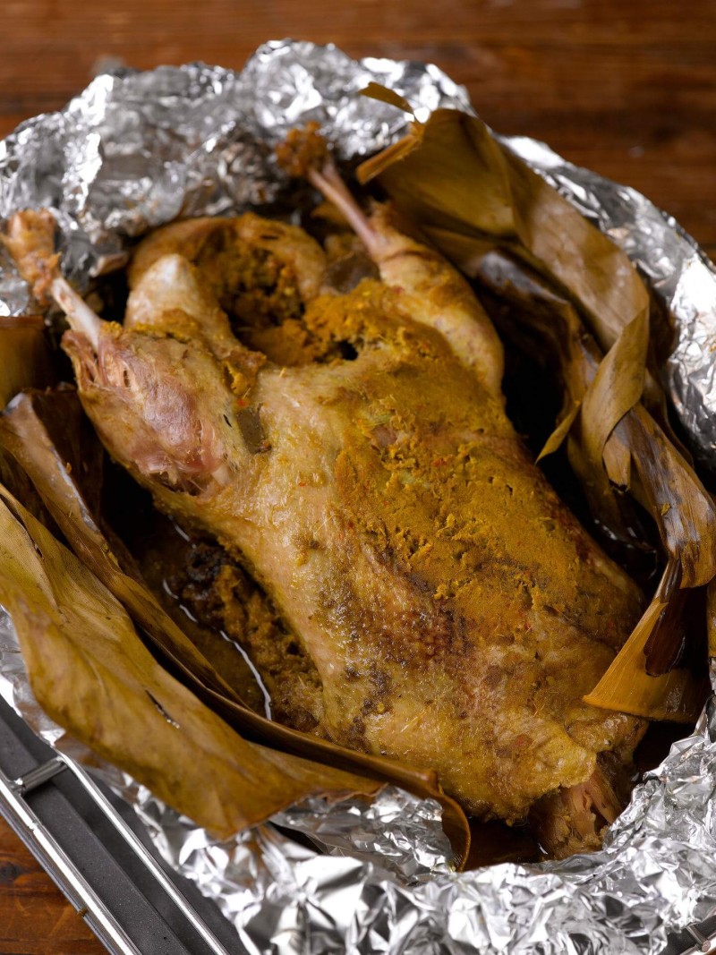 Whole duck stuffed with spices and wrapped in banana leaves, cooked in foil, resting in a tray.