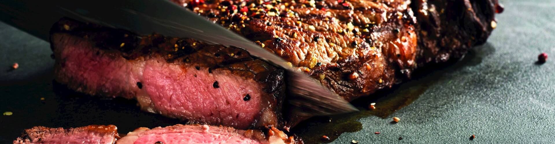 Primo piano di una bistecca di manzo perfettamente cotta che viene affettata, rivelando un interno rosa e succoso, cosparso di spezie su una superficie scura.