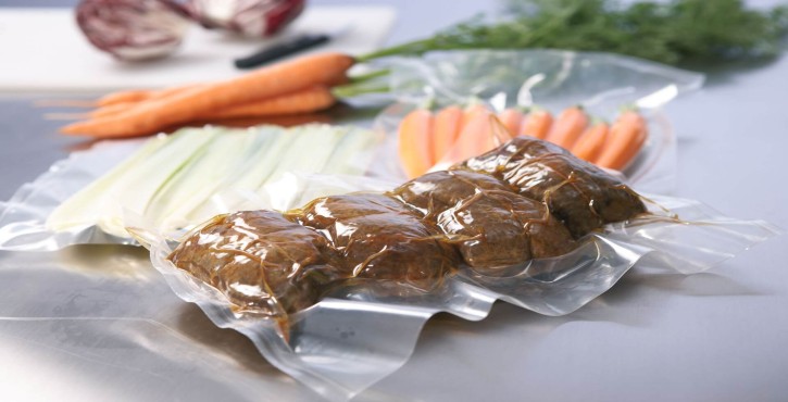 Vakuumverpackte Fleischrouladen neben frischen Karotten und Sellerie, alles auf einer Küchenarbeitsplatte arrangiert.