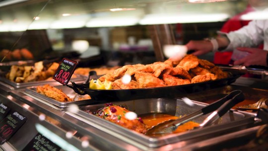 Un'affollata esposizione di cibo in un supermercato con pollo fritto, contorni e cartellini dei prezzi, con un membro del personale che serve i clienti.