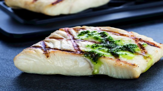 Grilovaný filet z halibuta s diagonálními grilovacími značkami, přelitý zelenou omáčkou, na černém servírovacím talíři.
