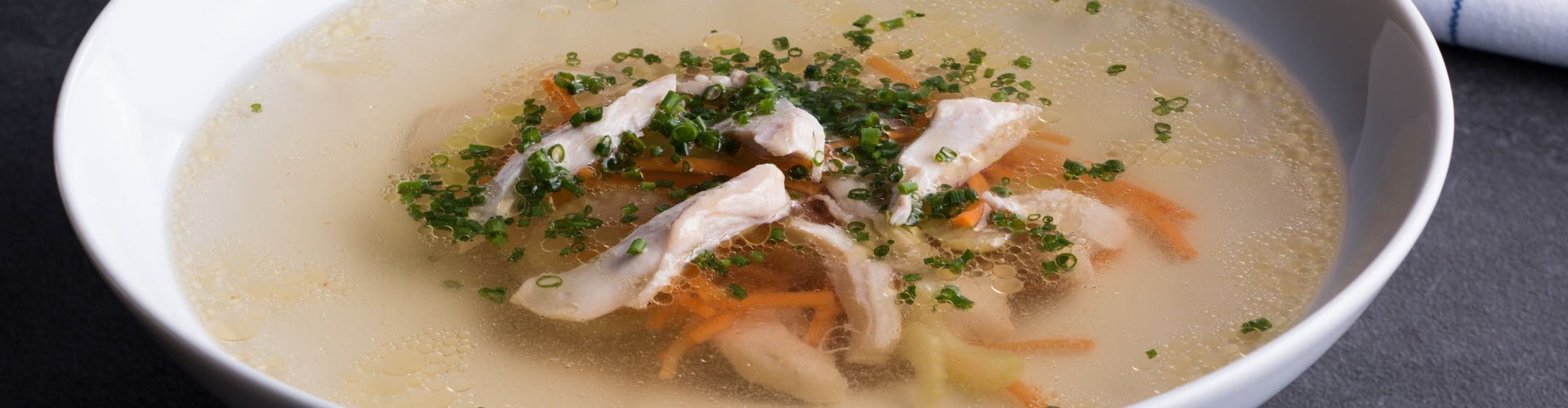 Eine Schüssel mit klarer Hühnerbrühe, gefüllt mit zerrissenem Hähnchen und buntem Gemüse, garniert mit Schnittlauch, auf einer dunklen Oberfläche.