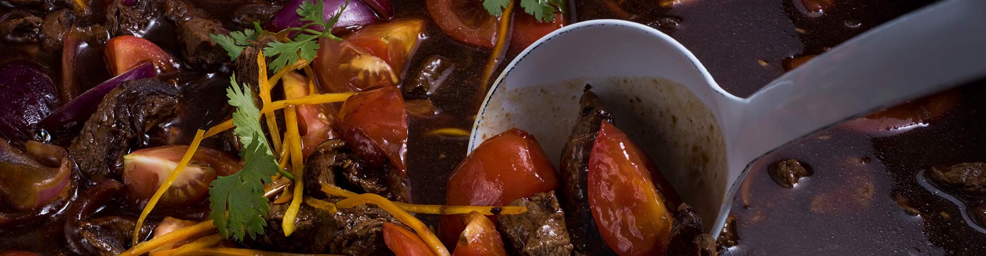 A close-up of a flavorful beef stir-fry dish with tomatoes, cilantro, and vibrant vegetables being served with a ladle.