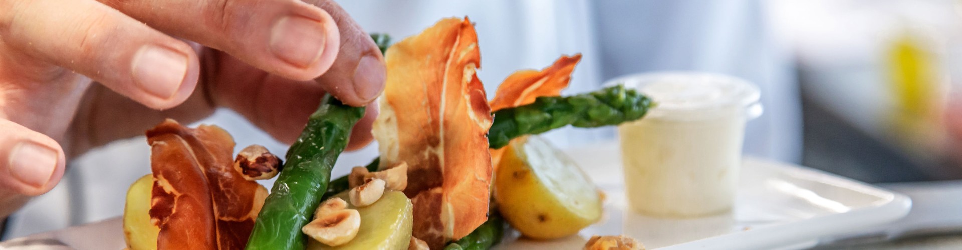 A hand garnishing a plate with asparagus, crispy prosciutto, baby potatoes, and hazelnuts, accompanied by a small container of sauce.