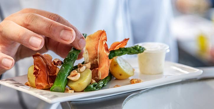  A chef plating asparagus, potatoes, and garnishes with a hand, along with a sauce on the side.