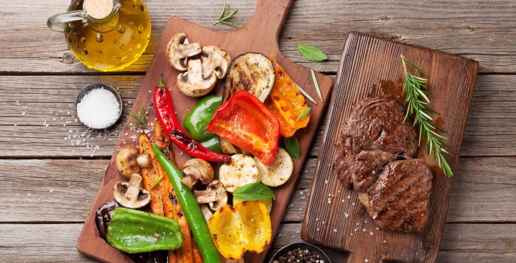 Légumes grillés (poivrons, champignons et carottes) sur une planche à découper, avec des steaks et de l'huile d'olive en arrière-plan.