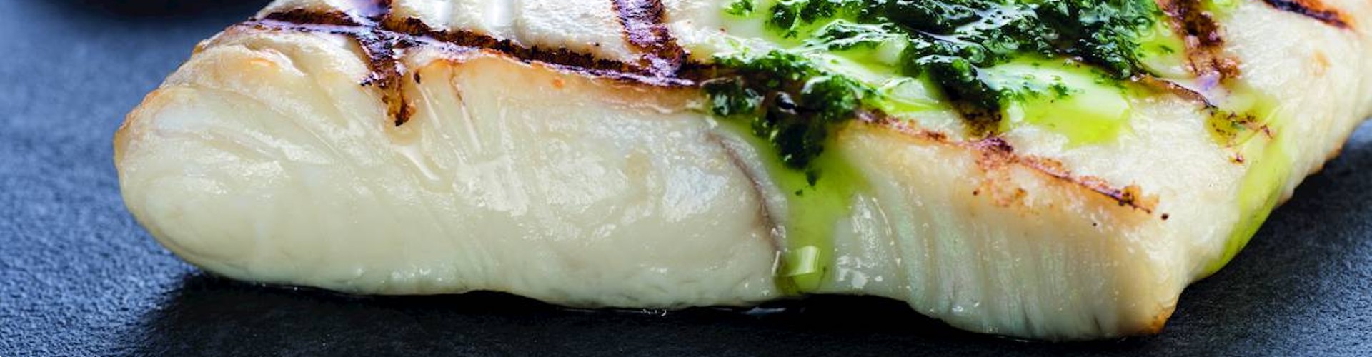 A grilled halibut fillet drizzled with green sauce, served on a dark surface.