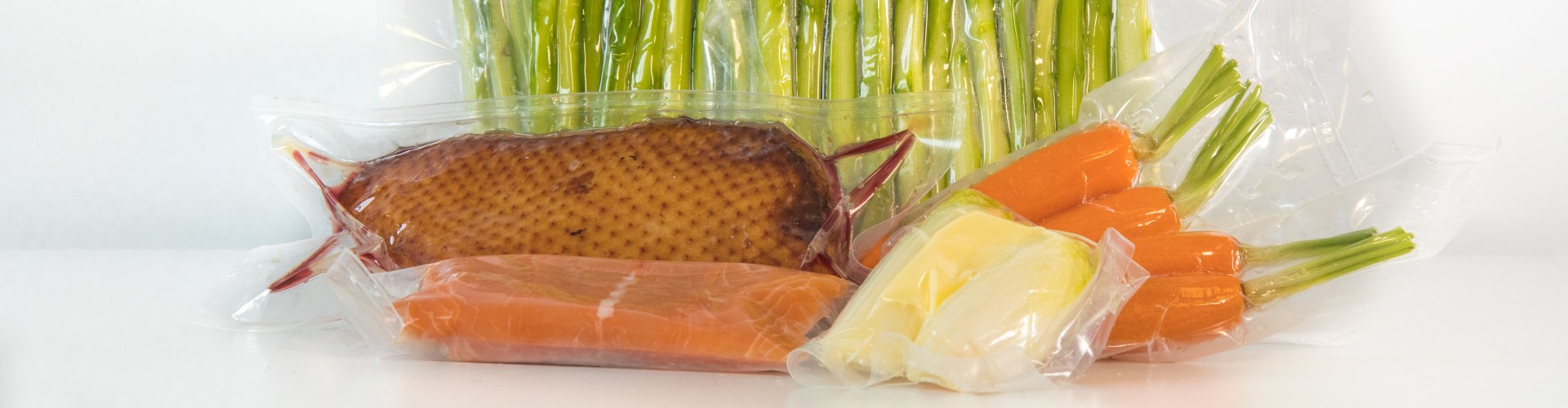 Various sous vide vacuum-sealed food items, including asparagus, salmon, smoked fish, and carrots, arranged on a white background.