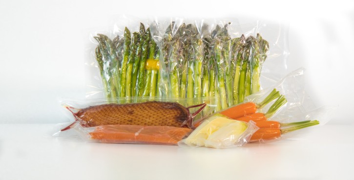 Diverses denrées alimentaires sous vide, dont des asperges, du saumon, du poisson fumé et des carottes, disposées sur un fond blanc.