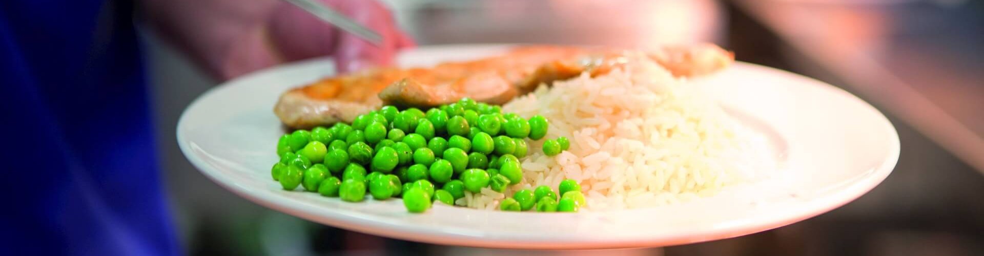 Gros plan d‘une assiette contenant du riz, des pois verts et un morceau de poulet dans une cafétéria, tenue par la main d‘une personne.