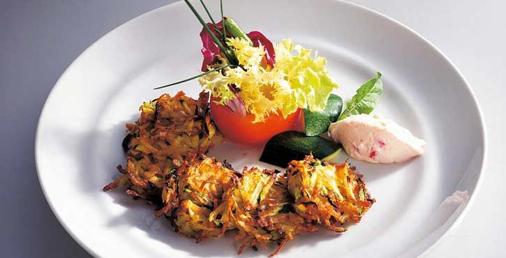 Ein Teller mit knusprigen Zucchini-Röstis, die kreisförmig angeordnet sind, garniert mit frischem Salat, Tomate und einem cremigen Dip.