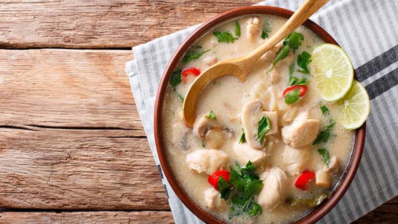 A bowl of Tom Kha Gai soup, featuring chicken, mushrooms, lime, and cilantro, served with a wooden spoon on a textured cloth.
