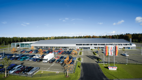 Aerial view of Rational's manufacturing plant, showcasing a modern facility with a large parking area filled with cars, surrounded by trees and blue skies.