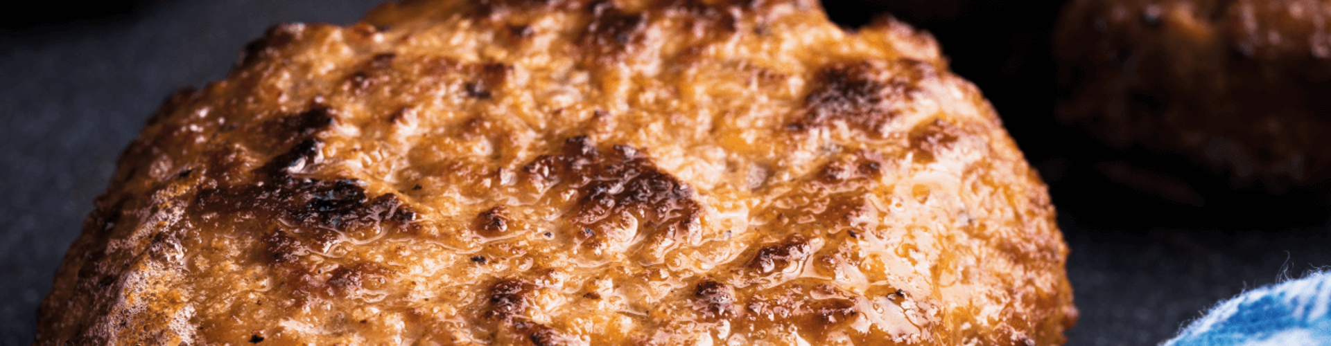 A close-up of a grilled hamburger patty with a crispy, golden-brown crust, glistening with juices, on a dark plate.