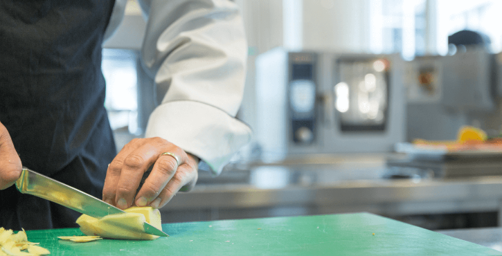 A chef prepares a potato on a green cutting board using a knife.