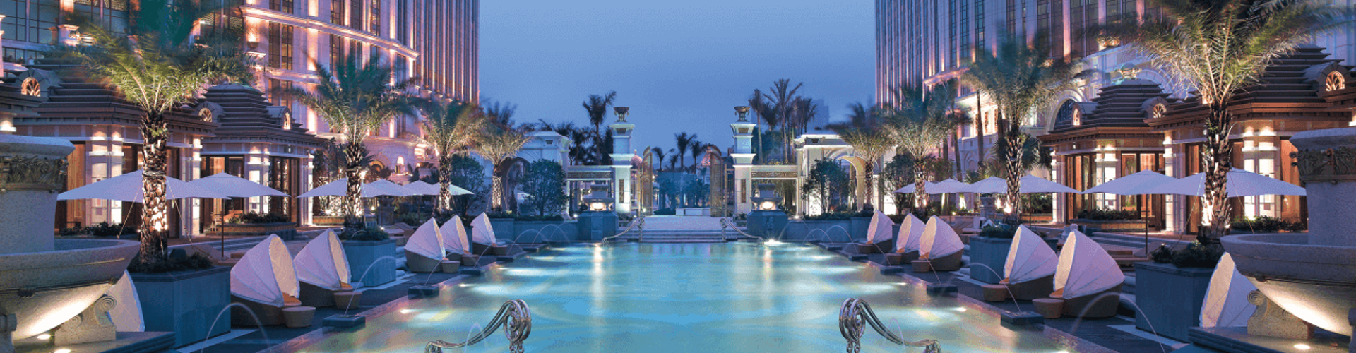A luxurious hotel with a large pool, cabanas, and palm trees illuminated at night.