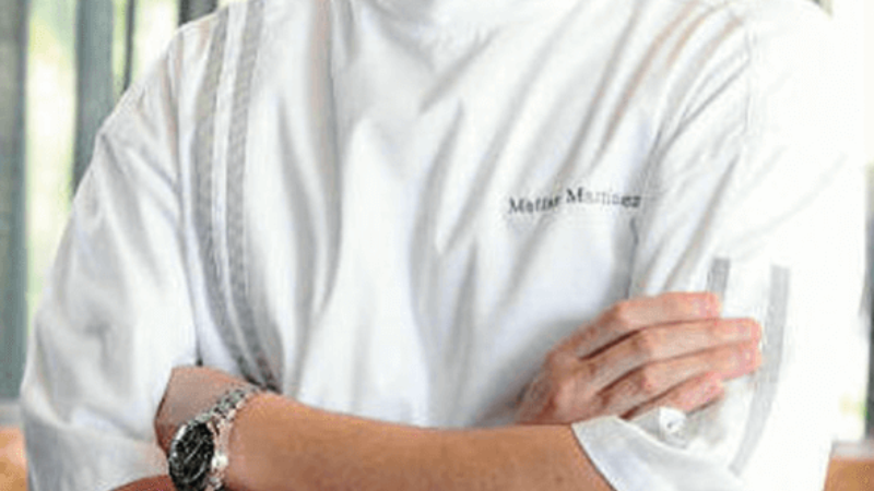 A chef standing with arms crossed, wearing a white chef‘s coat and glasses, with a curtain backdrop.