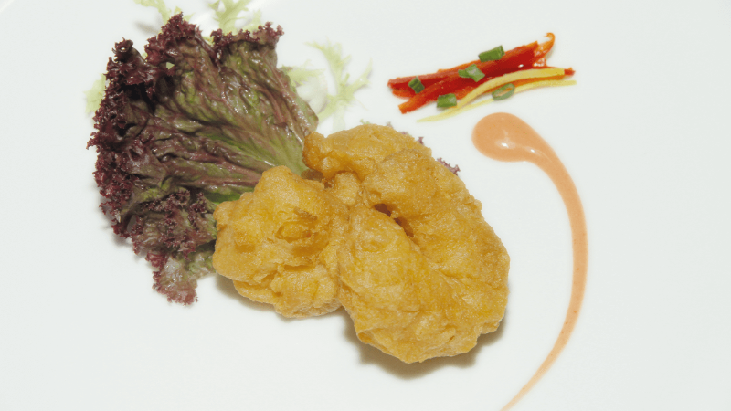 A fried dish with a golden crust, served with red lettuce, colorful sliced peppers, and a swirl of sauce.