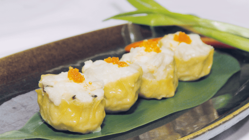A plate of dim sum dumplings with yellow wrappers, topped with orange roe, served on a leaf garnish.