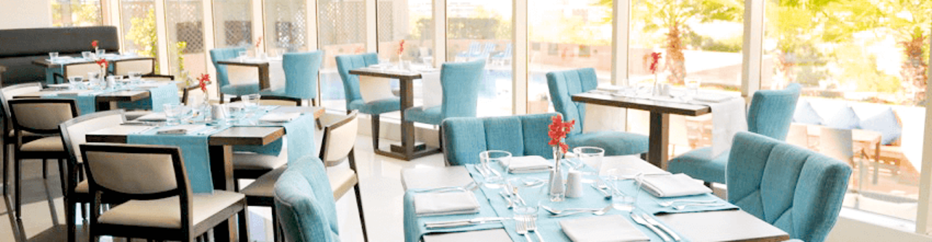 A bright, modern restaurant with turquoise chairs and tables set for dining, large windows offering a view of palm trees outside.