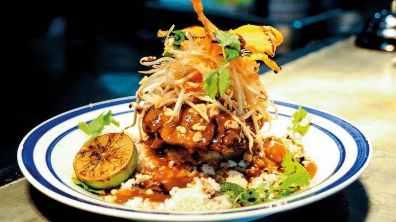 A beautifully plated dish featuring rice, a grilled protein topped with sprouts, herbs, and a lime slice.