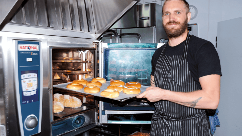 A baker in an apron holds a tray of freshly baked bagels in front of an oven.