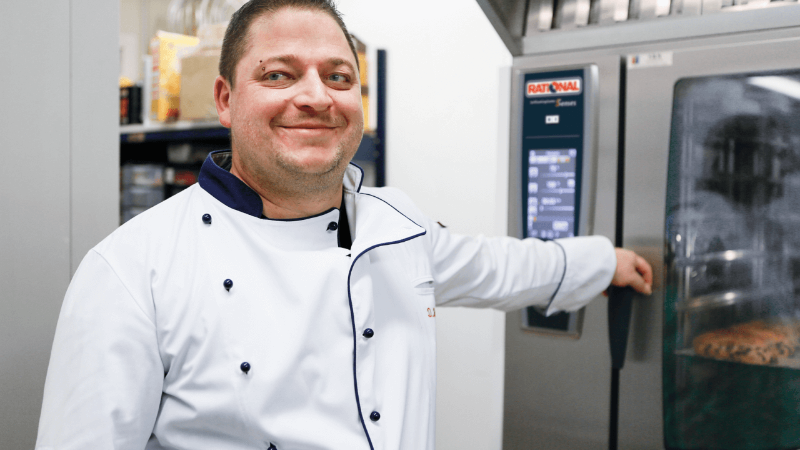 A smiling chef in a white coat stands next to a Rational oven, pointing at it while looking at the camera.
