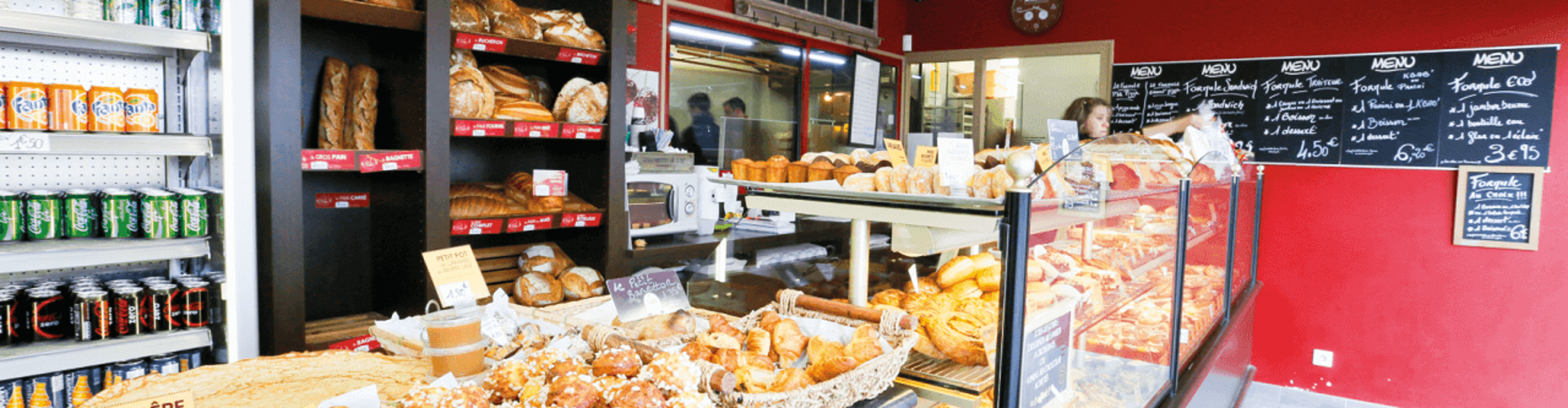 A bakery display featuring various breads, pastries, and baked goods, with a menu board on the wall and shelves of drinks on the left.
