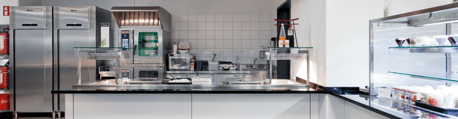 Interior view of a modern commercial kitchen with stainless steel appliances, including a ventilation hood, range, and refrigeration units. 