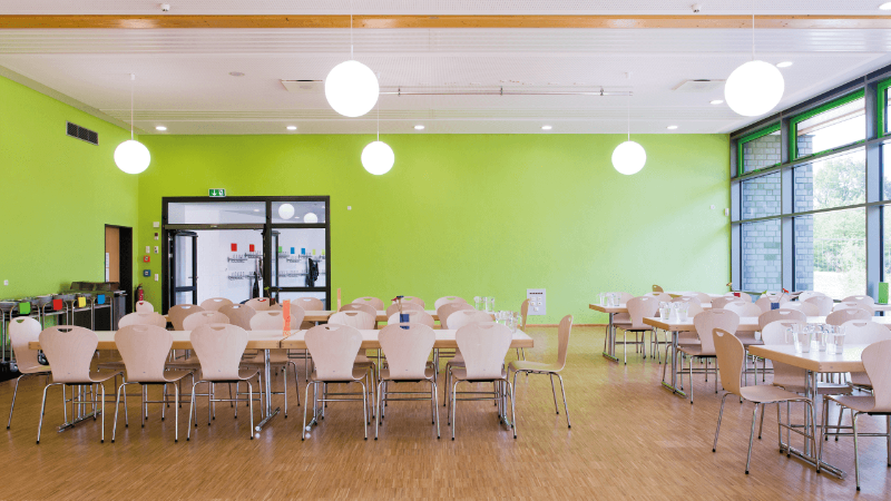 Spacious cafeteria with vibrant green walls and round tables set up with chairs. The room features large windows allowing natural light, and modern, globe-style hanging lights.
