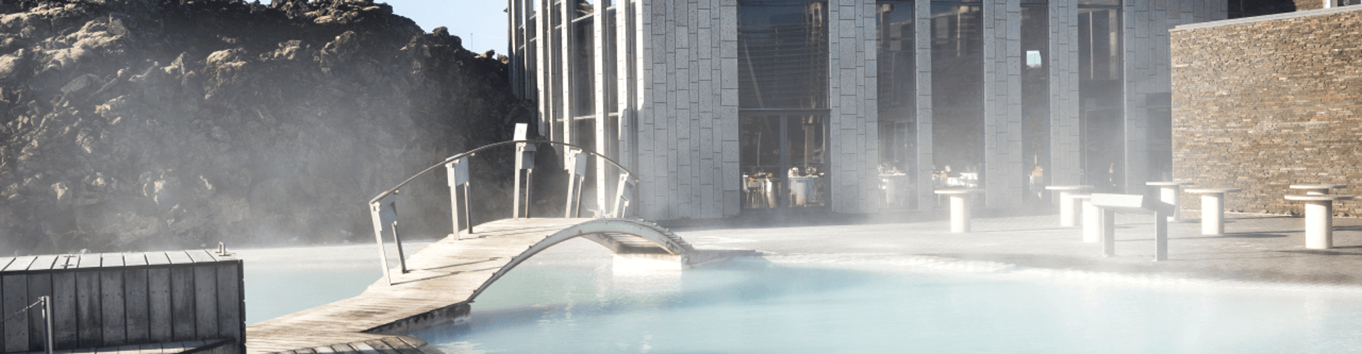 A modern spa with a wooden bridge leading over a steaming blue pool, surrounded by rocky terrain and a sleek building.