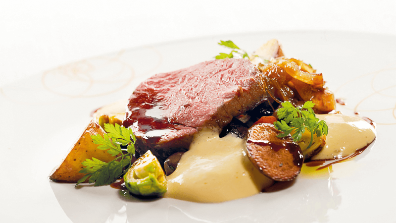 A plate of sliced beef with roasted vegetables, Brussels sprouts, carrots, and creamy sauce, garnished with herbs.