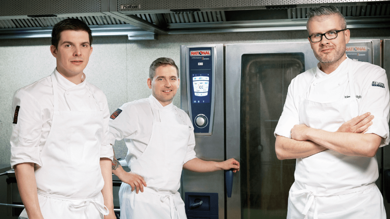 Three chefs in white uniforms stand together in a kitchen, next to a Rational oven, with a modern kitchen backdrop.