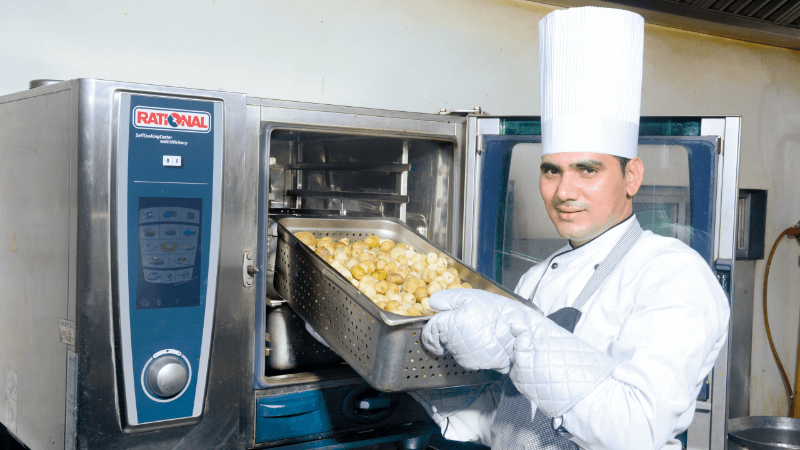 A chef wearing a white uniform and gloves, holding a tray of food in front of a Rational oven.